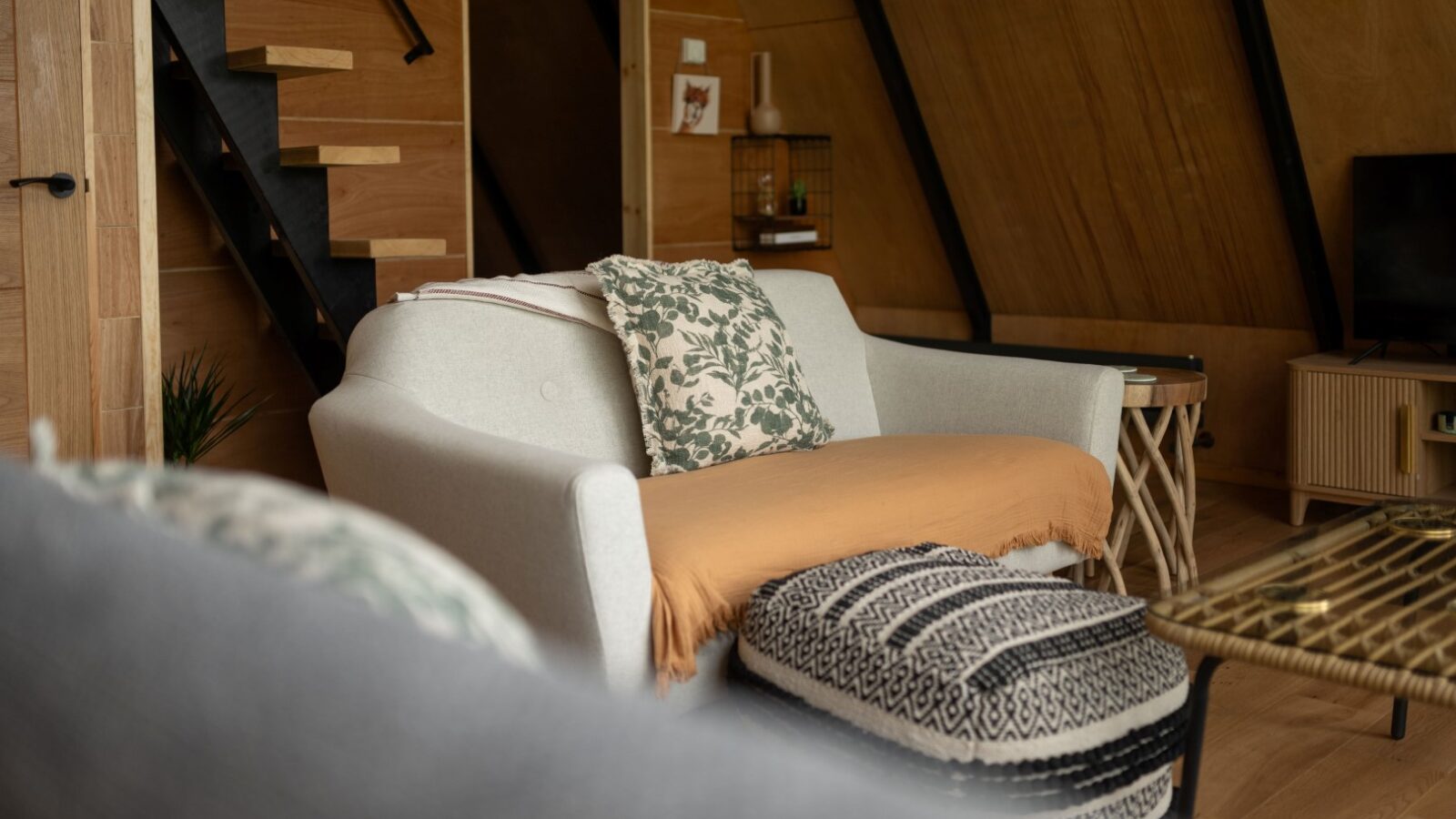 A cozy living room in Sigrid Lodge features a white armchair adorned with a floral cushion and an orangish-brown throw. In front, there is a patterned ottoman, a plant, and a modern staircase with wooden steps leading to an upper level. The room is warmly lit with wooden accents.