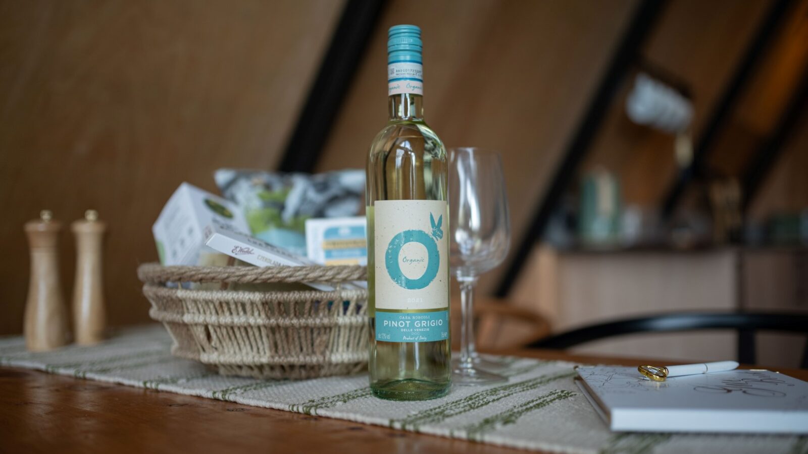 A bottle of Pinot Grigio wine sits on a wooden table in the cozy lodge, accompanied by a wicker basket filled with a small box and other items. Next to the wine bottle is an empty wine glass, ready for pouring. The background hints at blurred elements of Sigrid's charming kitchen or dining area.