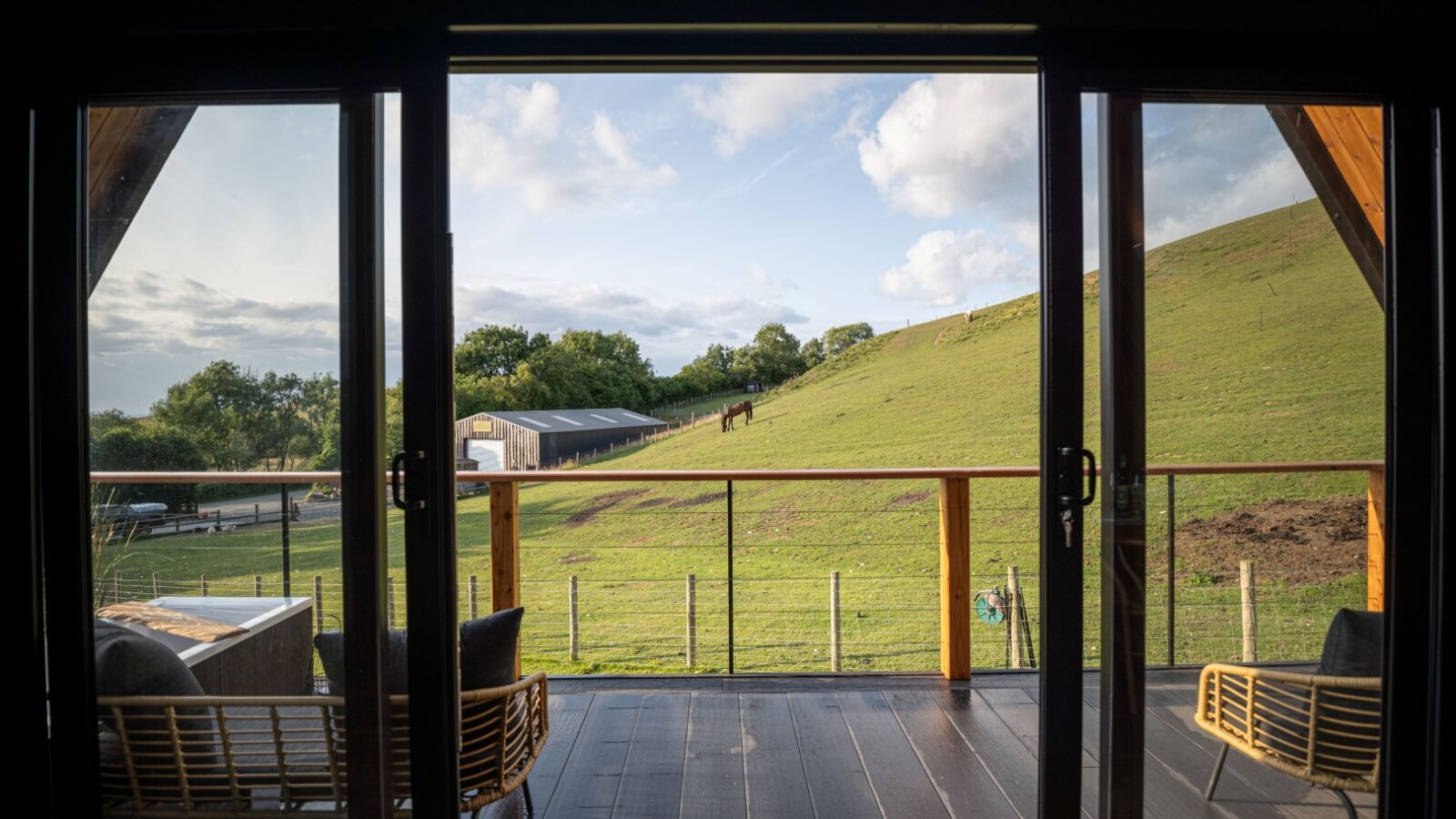 A serene countryside scene viewed through the sliding glass doors of Sigrid Lodge. Outside, a green hilly pasture with a lone horse is seen under a partly cloudy sky. The patio features modern outdoor furniture, including wicker chairs and a table, providing a cozy and inviting atmosphere.