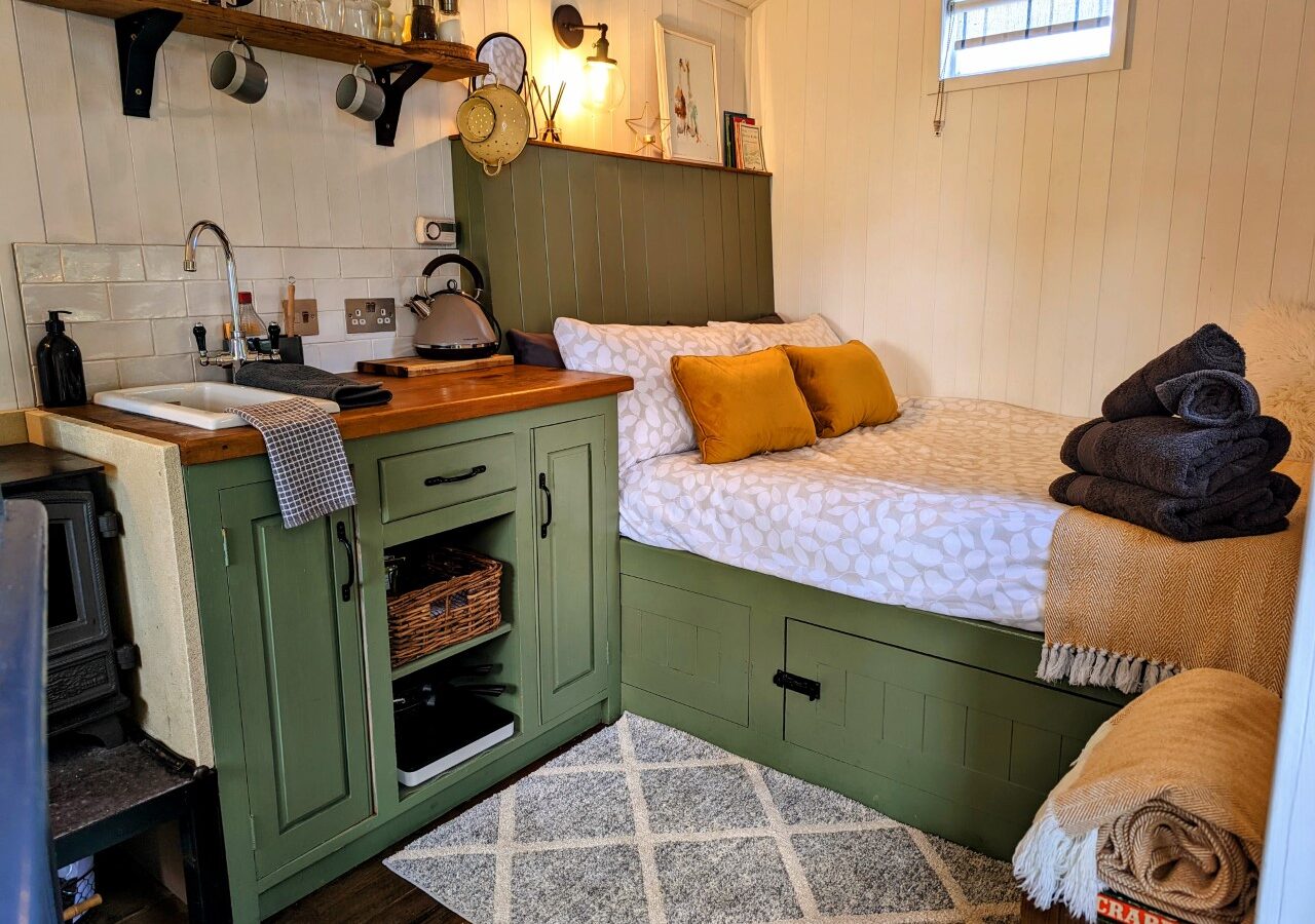 A cozy bedroom in a Shepherds Hut features a bed with mustard pillows and folded blankets. There's a green kitchenette with a sink and stove, wooden shelves with dishes, and a small rug near the wood-burning stove. The white walls provide a charming contrast to this rustic haven.