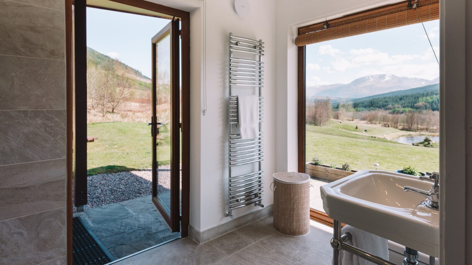 The bathroom, reminiscent of a scene from Frances Sketch Pad, boasts open doors and a large window framing a scenic view of mountains, lush grassy landscapes, and a meandering river.