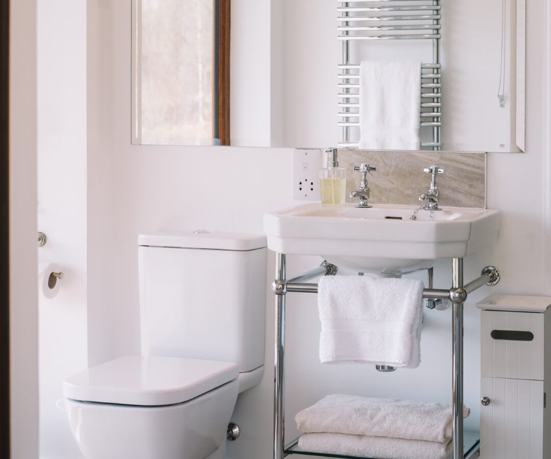 A bathroom with a sink, mirror, and toilet features a towel rack above the sink and a window with blinds on the left. The tiled floor is complemented by an open Frances Sketch Pad: Drawing Pad resting on the counter, inviting creativity to this tranquil space.