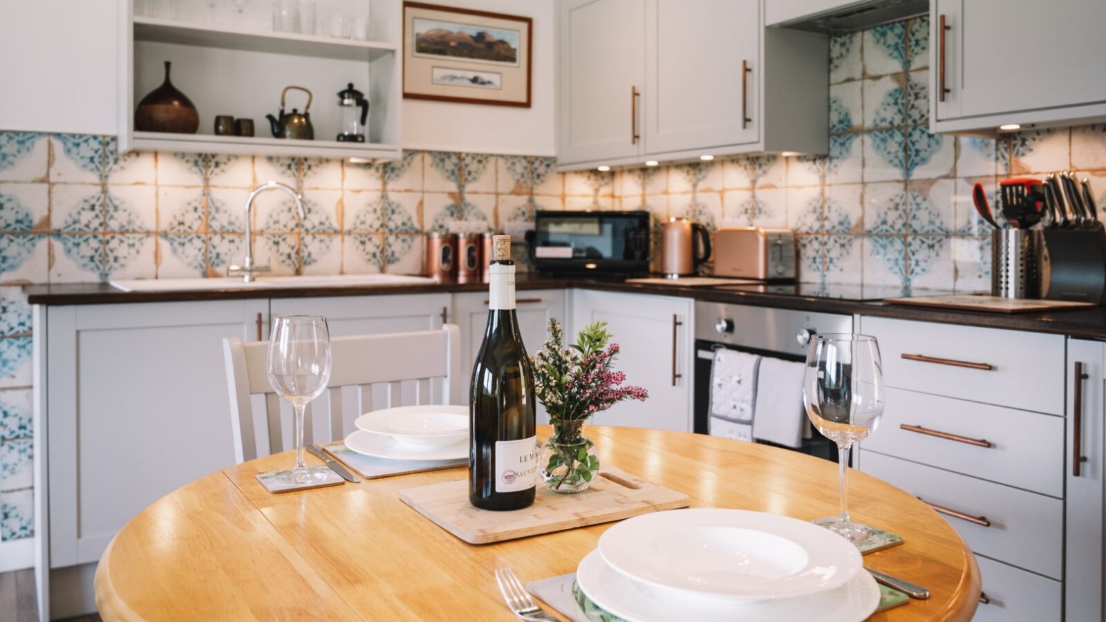 A cozy kitchen with a wooden table set for two, featuring plates, wine glasses, and a wine bottle. Frances sits at the table sketching on her pad while the tile backsplash adds an elegant touch in the background.