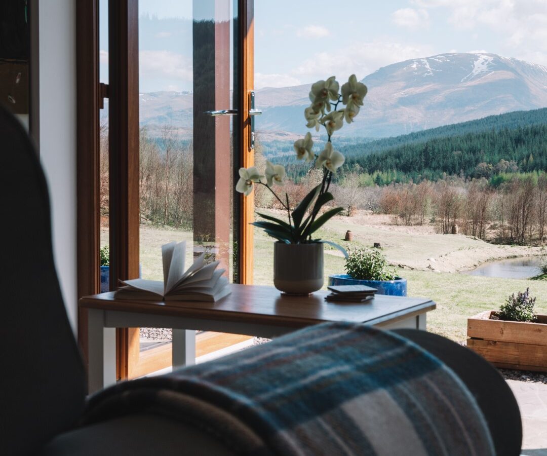 Open door revealing a mountain view; cozy chair with a plaid blanket and a table with Frances's sketch pad, alongside a potted plant and book.