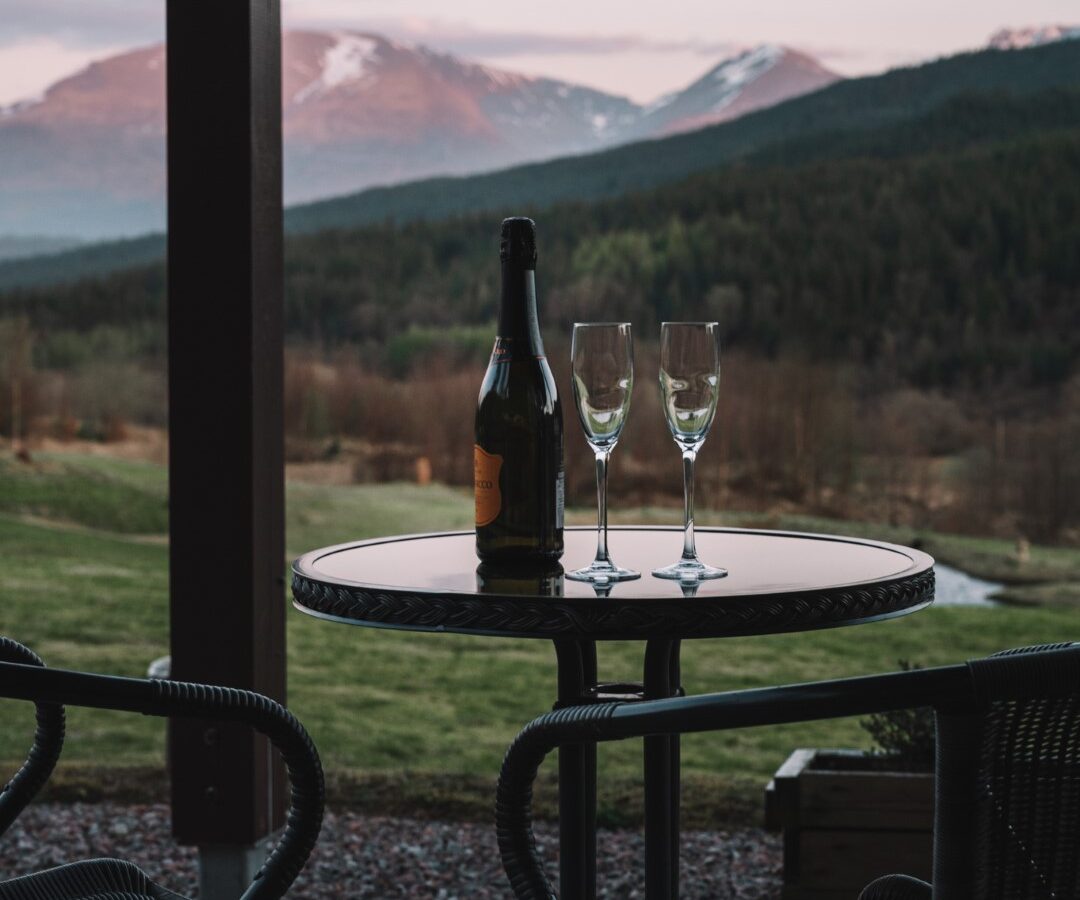 A sketch pad rests on the table beside a wine bottle and two glasses, capturing the stunning mountain landscape at sunset.