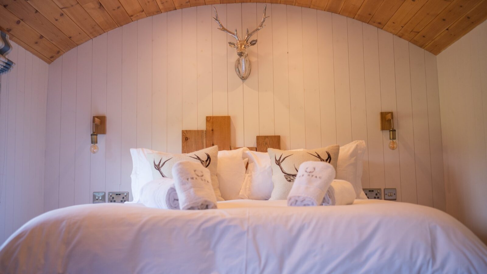 Cozy bedroom in The Vale with a wooden ceiling, white bedding, and deer antler decor on the wall, featuring two rolled towels on the bed.