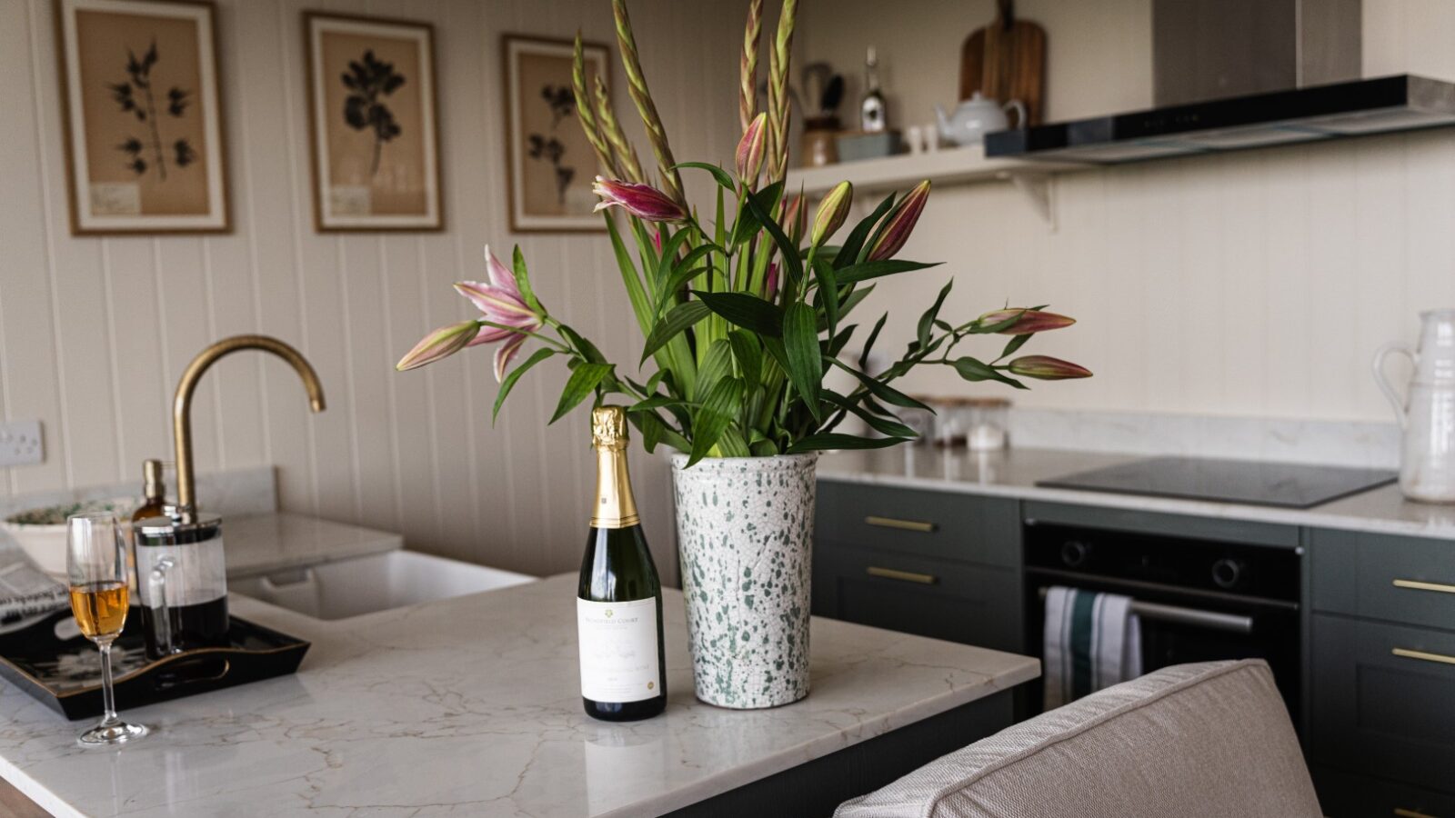 A modern kitchen with white countertops, a green potted plant, and a bottle of champagne captures the essence of luxury lodges. Framed art graces the wall, adding an elegant touch reminiscent of stars and vines.