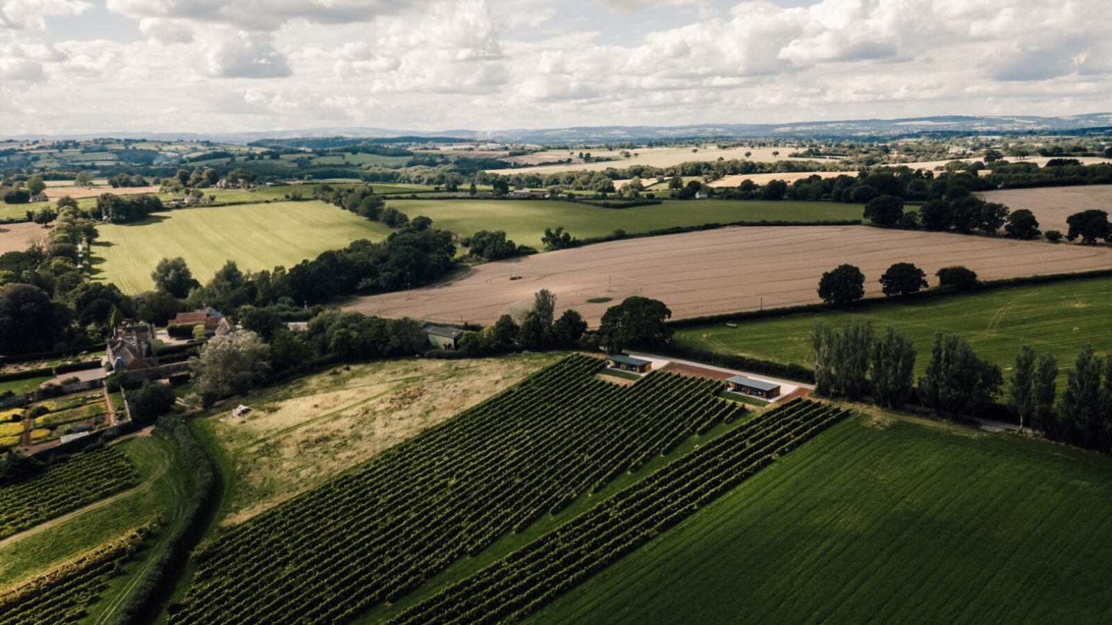 Aerial view of a rural landscape with fields, vineyards, and scattered trees under a partly cloudy sky, where luxury lodges nestle harmoniously, offering serene retreats amid the stars and vines.