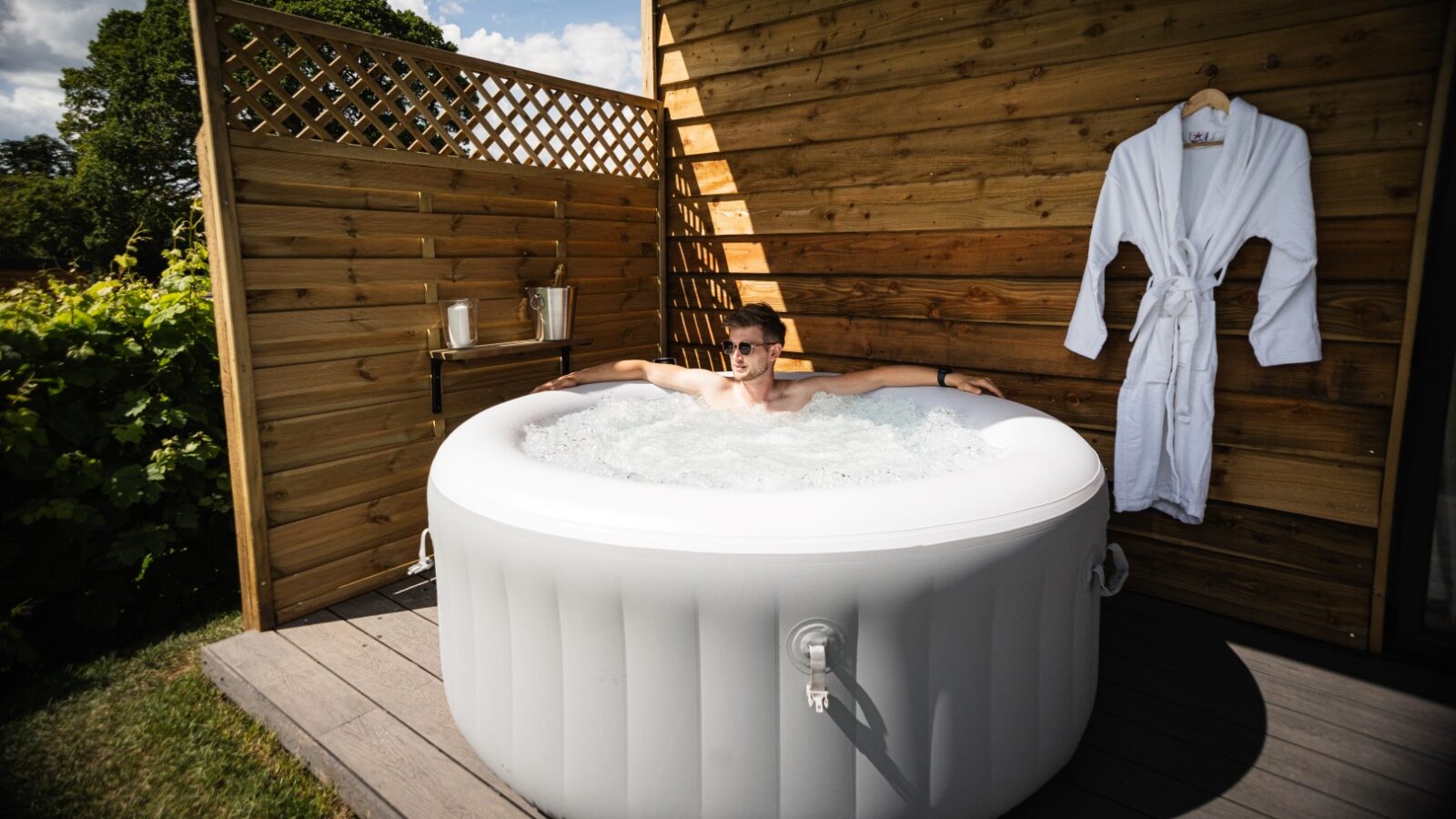 A person relaxes in a round inflatable hot tub outdoors at a luxury lodge. The setting features a wooden privacy screen, a hanging bathrobe nearby, and an enchanting view of stars and vines above.