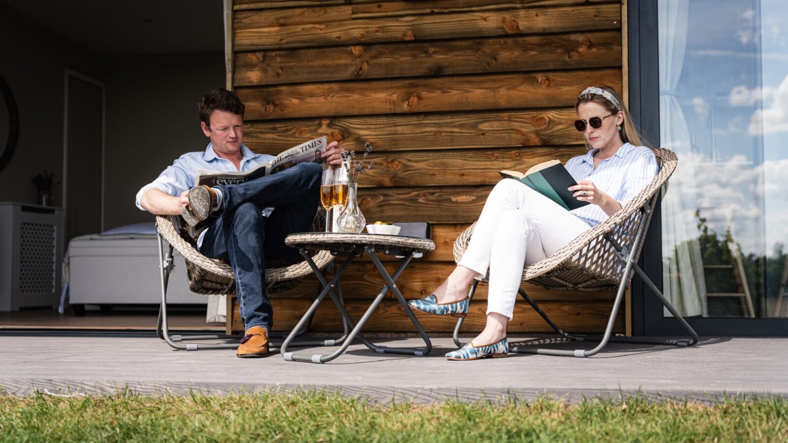 A man and woman sit on wicker chairs outside a wooden house, enjoying the serenity of their luxury lodge. He peruses a newspaper while she escapes into the pages of her book. Drinks rest on the table, framed by stars and vines that paint an idyllic backdrop.