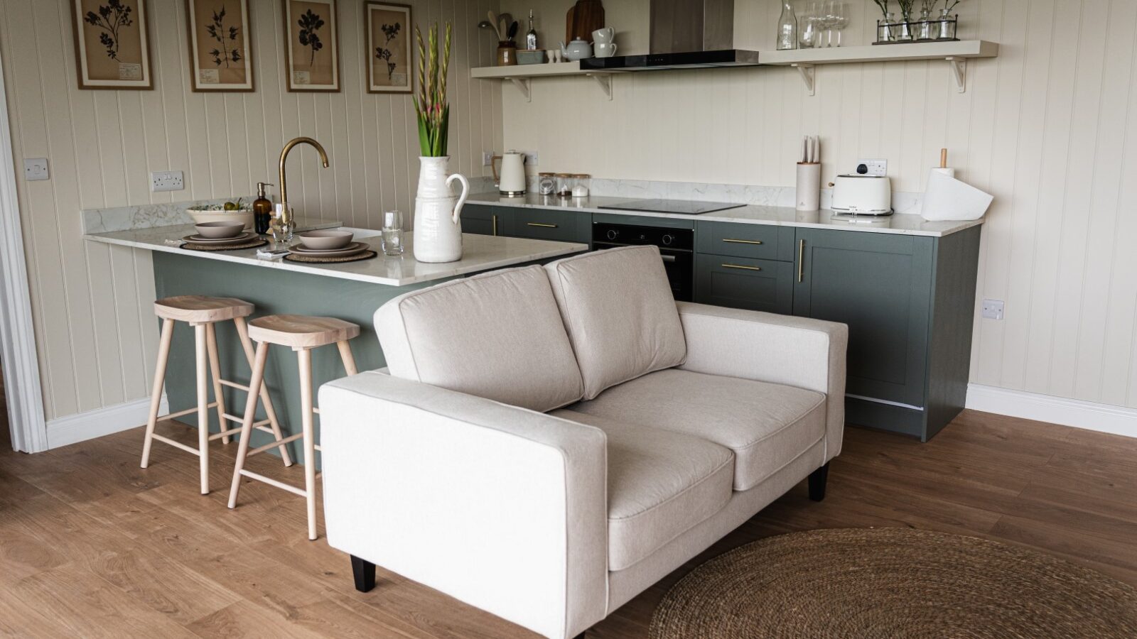 A modern kitchen and living area in luxury lodges, featuring a comfortable sofa, stylish bar stools, and a sleek countertop. Shelves display kitchenware while framed art decorates the walls.