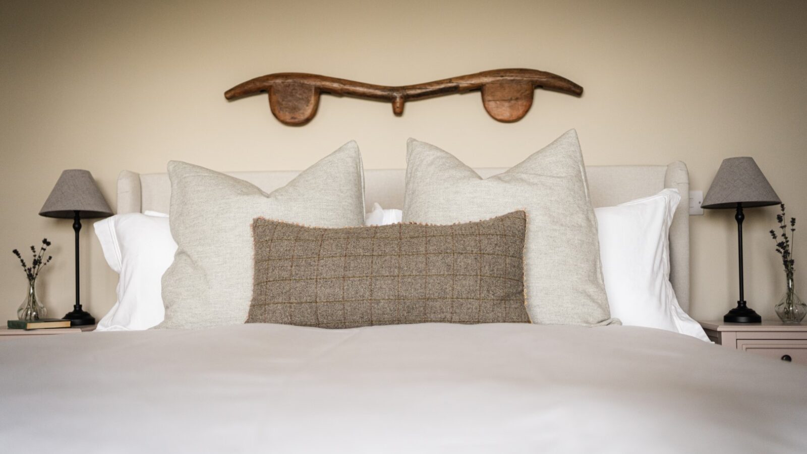 A neatly made bed with various pillows is centered under a wall decoration adorned with vines, flanked by two bedside tables with lamps.