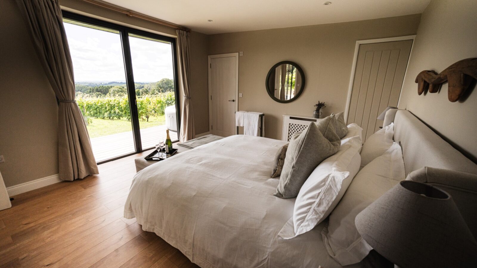 A luxurious bedroom featuring a large bed, round mirror, and open curtains that reveal the enchanting view of a vineyard outside. The elegant decor embodies neutral tones, creating a serene atmosphere. This is one of many luxury accommodations where stars and vines create an unforgettable experience.