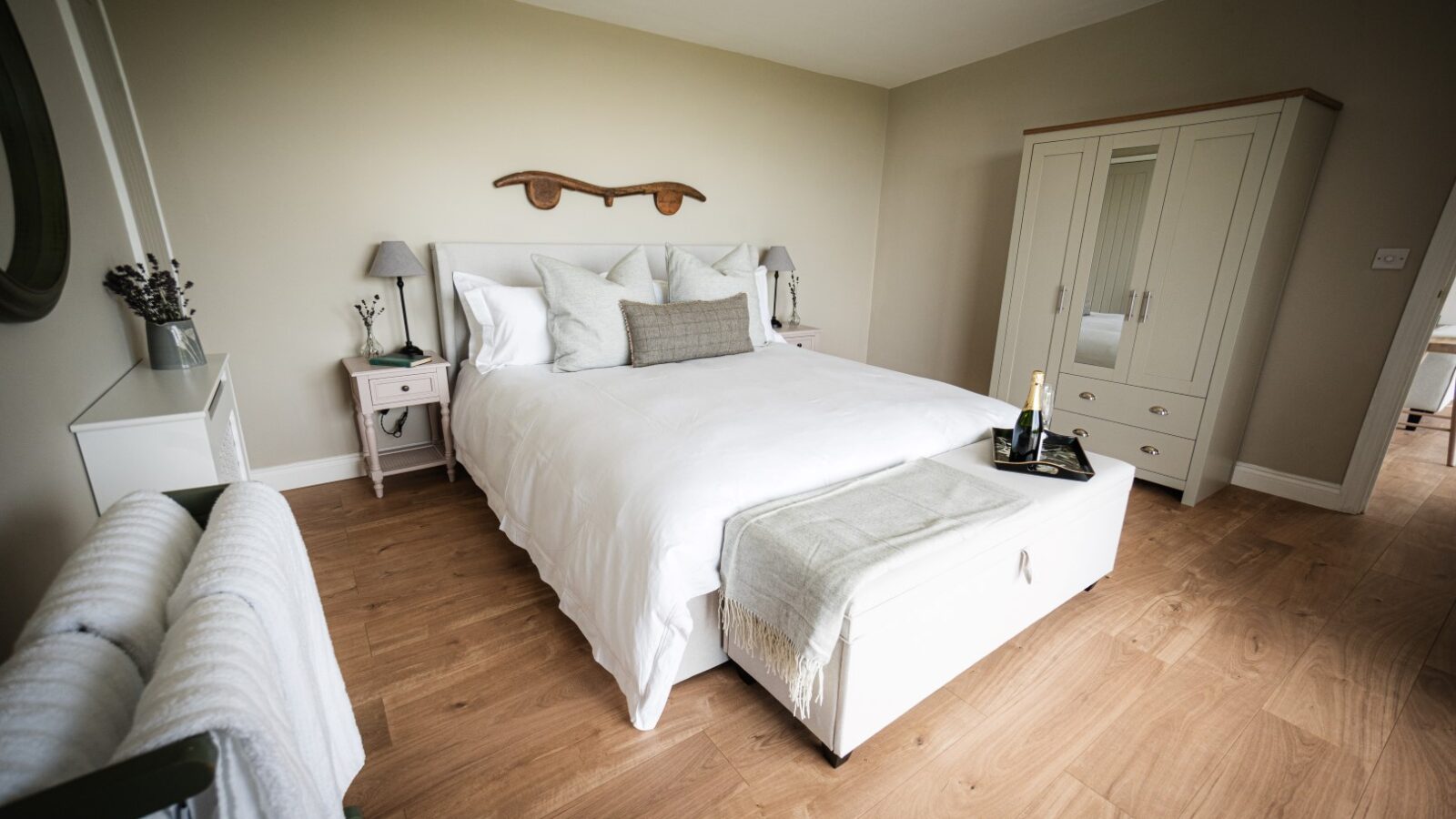 Experience luxury accommodations in a serene bedroom featuring a double bed adorned with white bedding. The elegant wooden floor complements the pristine white wardrobe, while on the bed, a tray elegantly showcases a bottle and glass, inviting relaxation amid stars and vines.