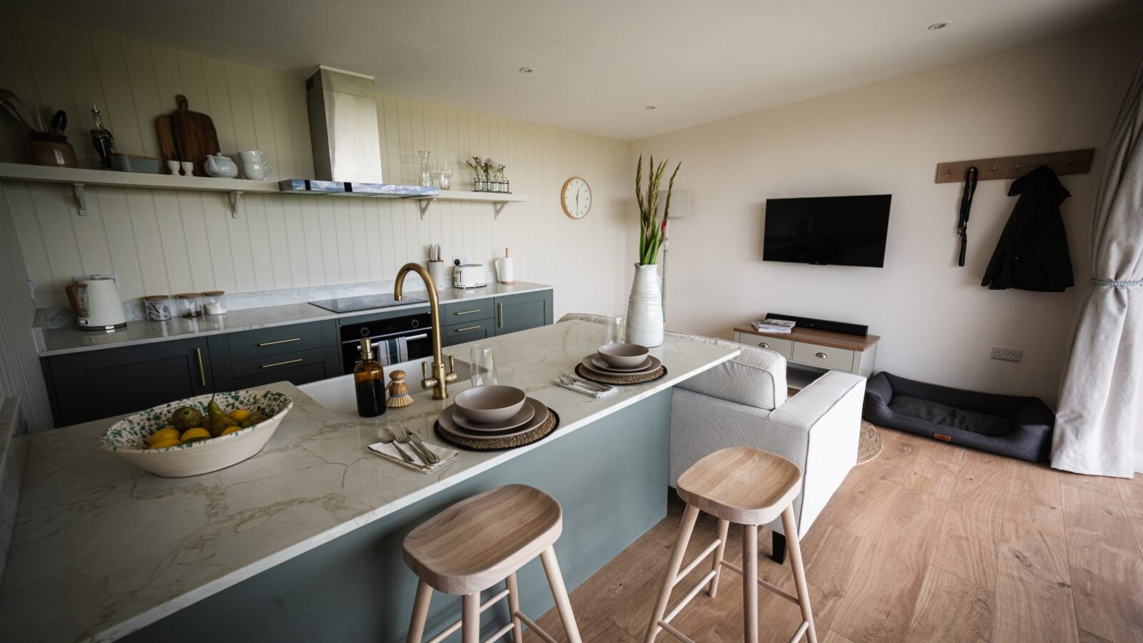 This luxury lodge features a modern kitchen with a marble island, sleek stools, and a set table. The living area offers comfort with its sofa and TV, while the corner is thoughtfully equipped with a dog bed for your furry companion—a perfect blend of stars and vines ambiance.