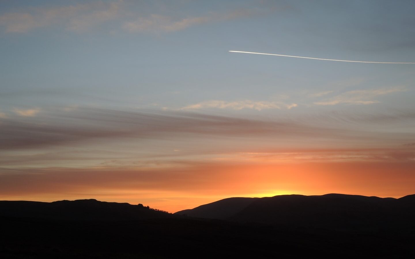 A serene sunset scene with the sun setting behind distant dark hills. The sky is painted with gradients of orange, pink, and blue, while a contrail from an aircraft crosses the upper part of the sky. Nearby, a cozy holiday cottage completes this tranquil atmosphere reminiscent of scenes from Lambing Live.