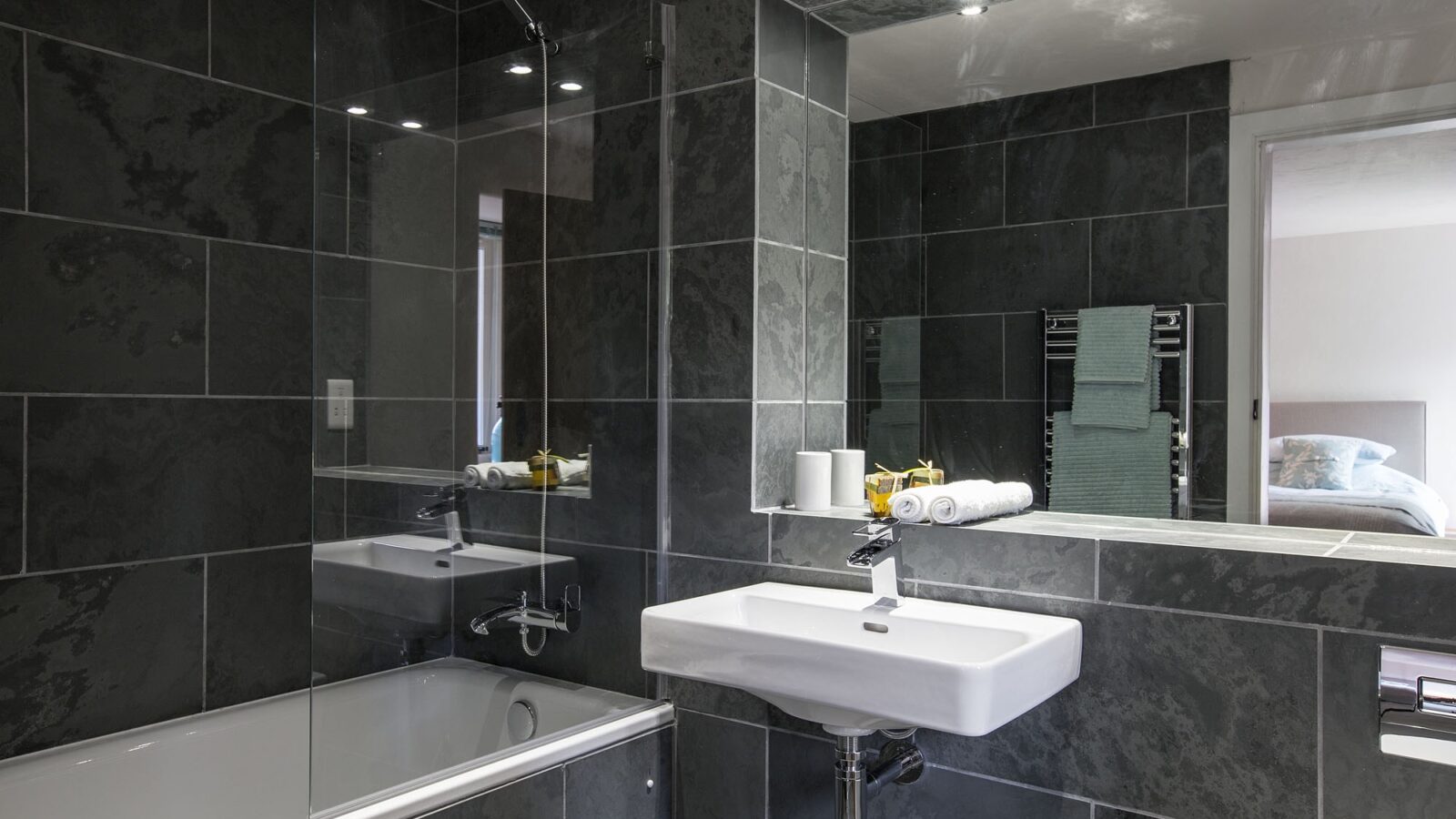 A modern bathroom with dark grey marble tile walls and floor. It features a white rectangular sink with a mirror above it, a bathtub with an overhead shower, and a wall-mounted towel rack with green towels. The bedroom is visible through the open doorway. Perfect for your stay at our holiday cottage.