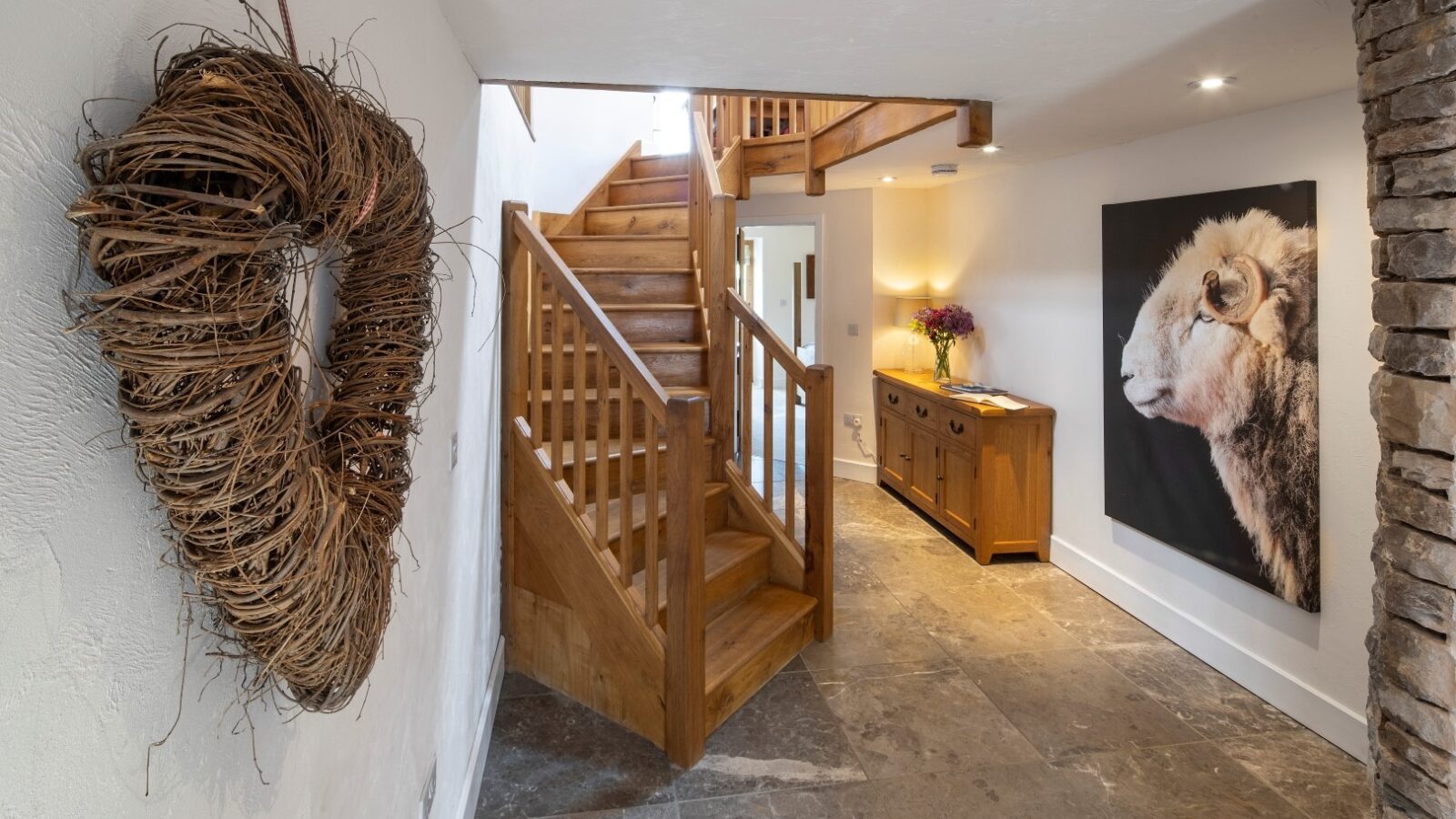 Bright entryway with wooden staircase, stone floor, and wreath on the left wall. A large sheep painting and a wooden dresser with a vase of flowers are on the right. Warm lighting enhances the welcoming atmosphere of this charming holiday cottage.