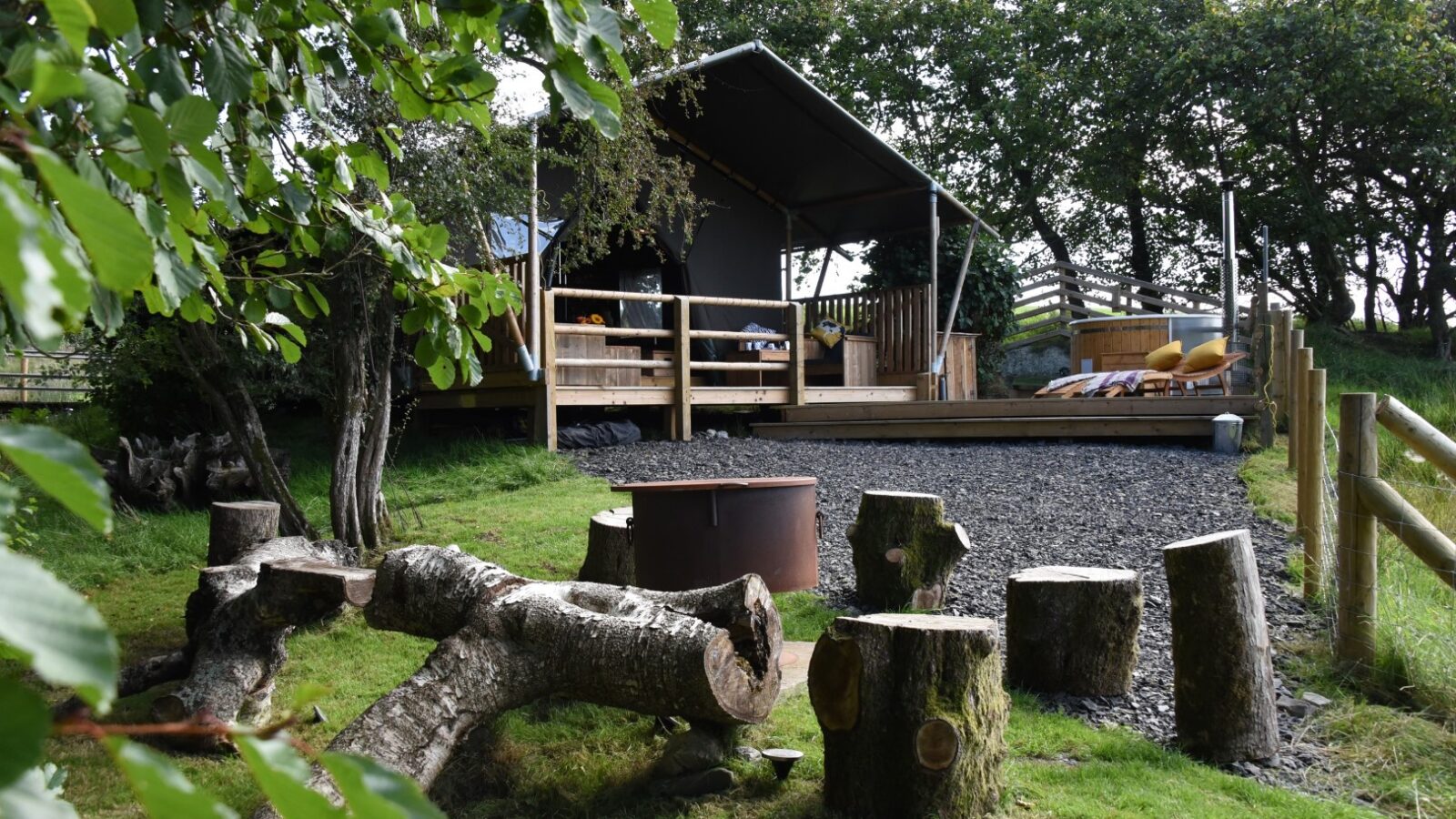 A rustic cabin with a wooden deck and outdoor seating is surrounded by trees, reminiscent of a serene lodge getaway. With chopped logs and a fire pit in the foreground, it offers an experience akin to a stellar safari adventure.