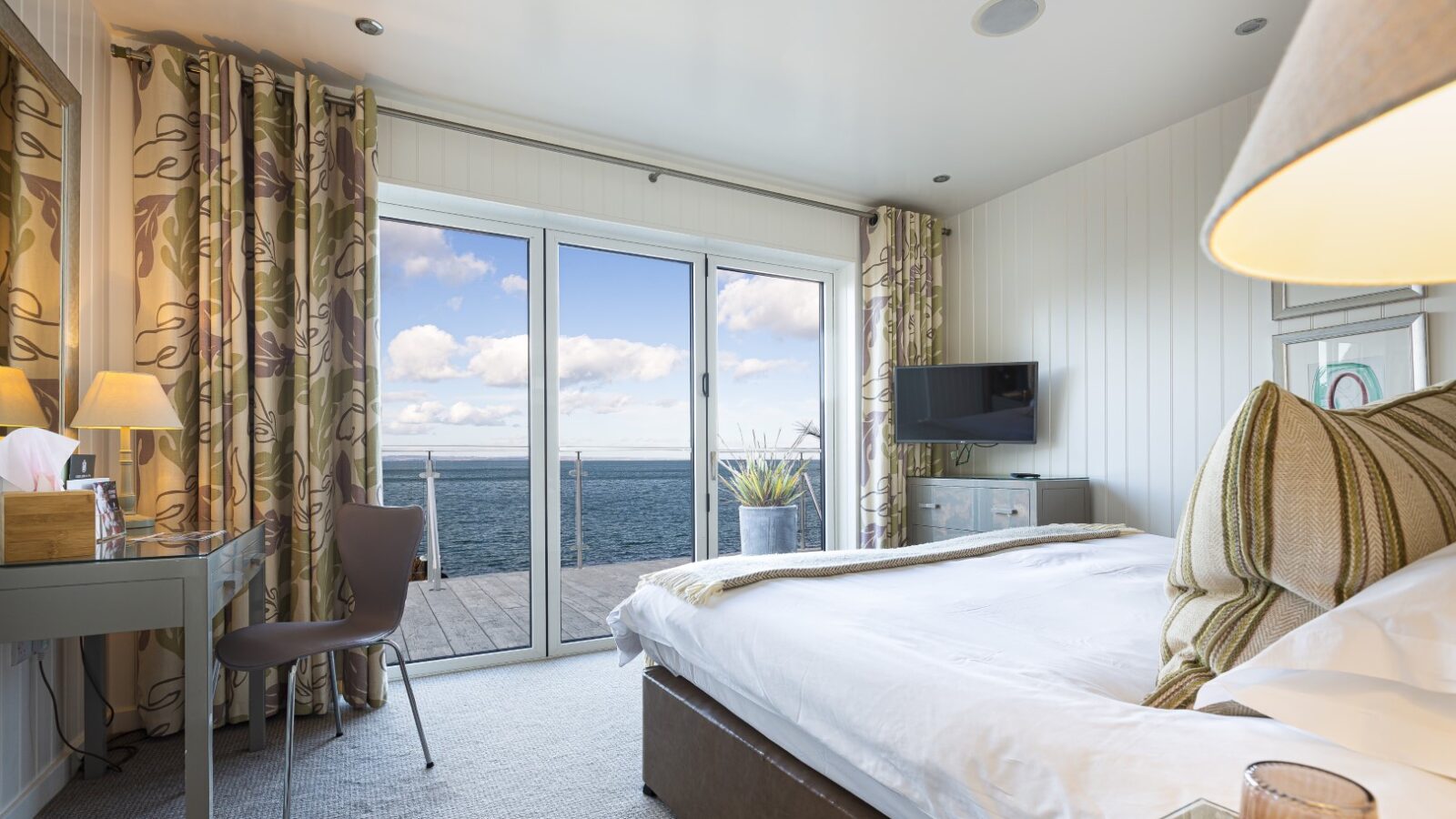 The bedroom, reminiscent of elegant suites, boasts a large bed, a desk, and a chair. Glass doors open onto a balcony offering stunning sea views akin to those from charming beach huts.