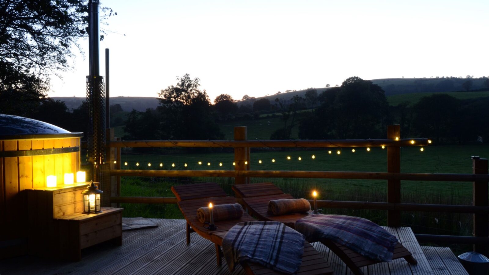 A wooden deck at sunset with two loungers, cozy blankets, glowing lanterns, and string lights offers a stellar view of the rural landscape, perfect for a lodge retreat.