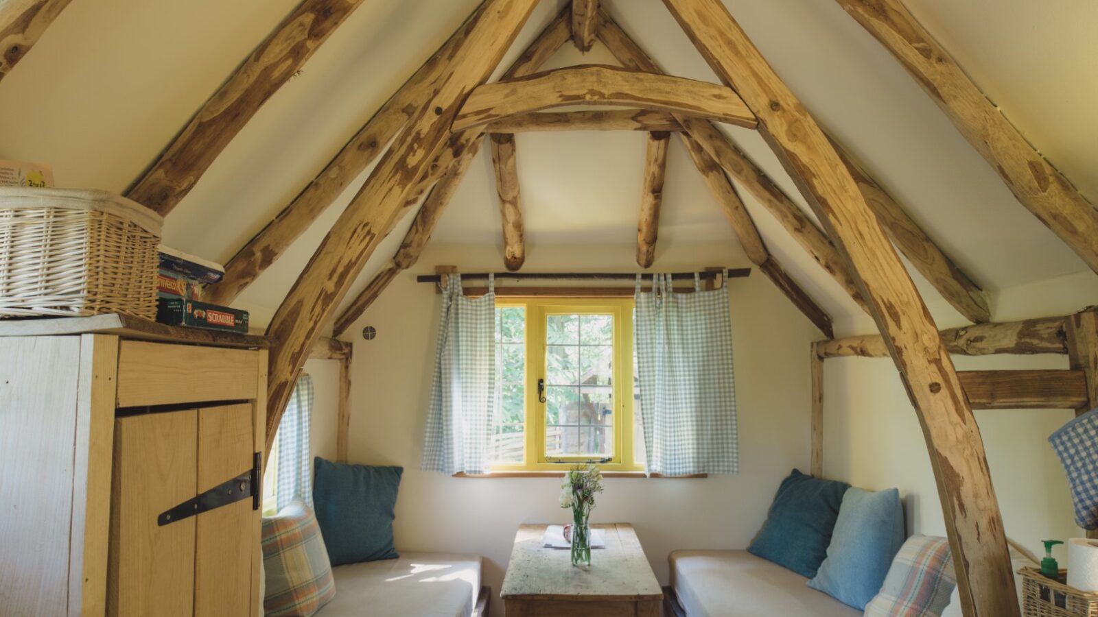 Rustic wooden cabin interior with two benches, a small table, and a window adorned with checkered curtains, reminiscent of a cozy woodcutter's retreat. A wicker basket rests on a shelf, adding to the charming cottage feel.