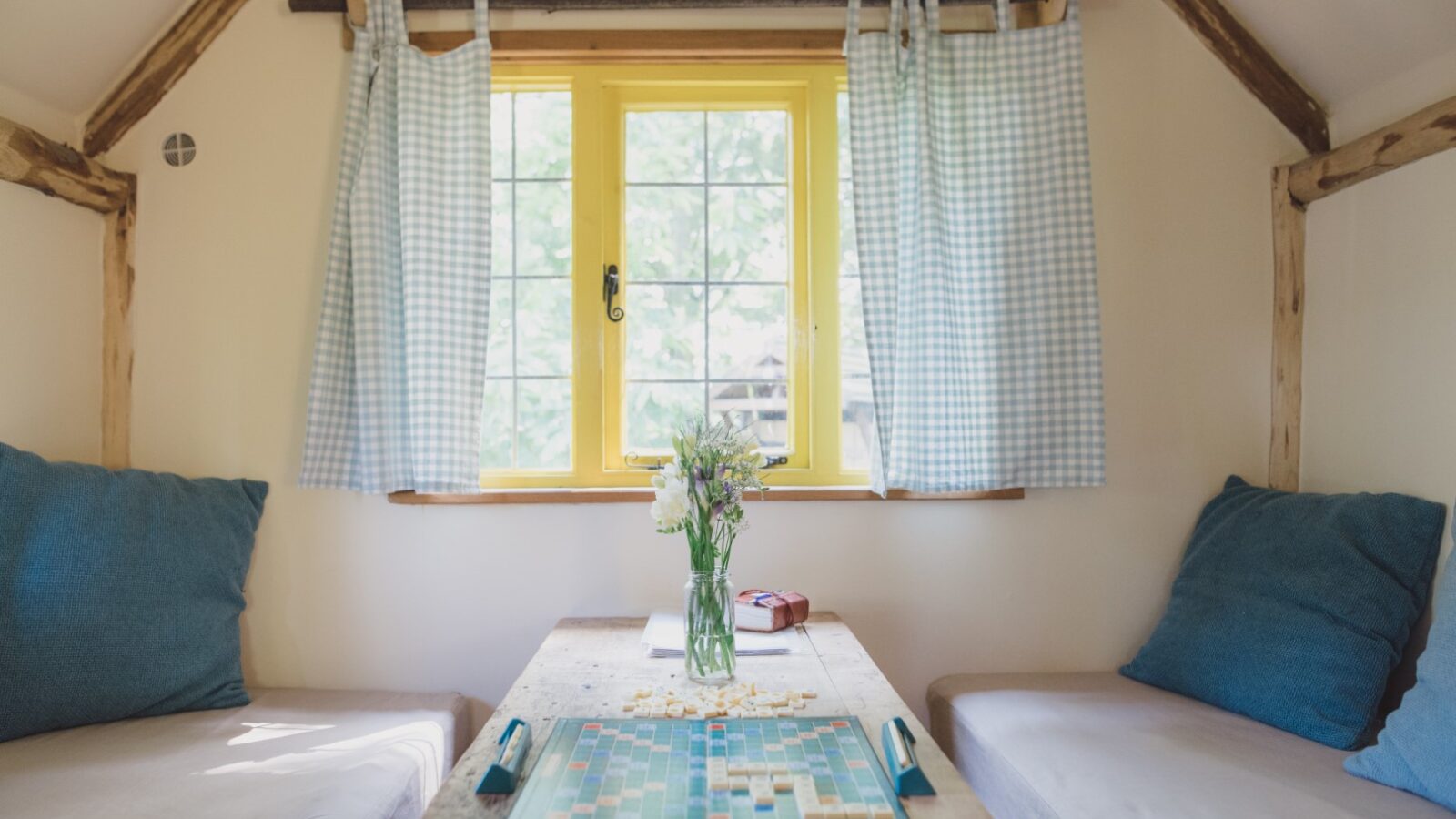 This small cozy room, reminiscent of a Woodcutters Cottage, features a wooden table, a flower vase, Scrabble game, blue cushions, and checkered curtains on a yellow window.