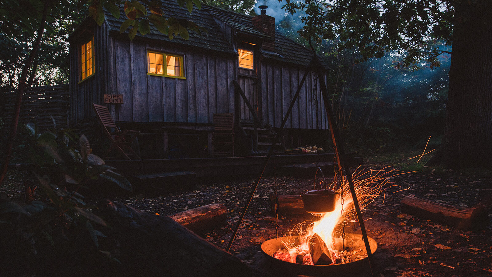 A small wooden cottage with lit windows nestled in a forested area, where woodcutters often gather. In the foreground, a tripod holds a pot over a crackling campfire, inviting warmth and camaraderie.