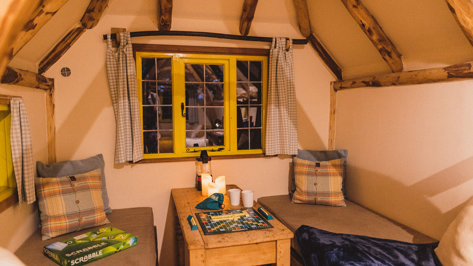 Cozy room in a quaint Woodcutters Cottage featuring a Scrabble board on a wooden table, two cushioned benches, plaid pillows, and a yellow-framed window.