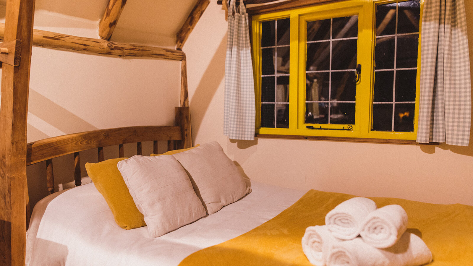 Cozy cottage bedroom with a wooden bed, yellow and white bedding, pillows, rolled towels, and a window adorned with checked curtains.