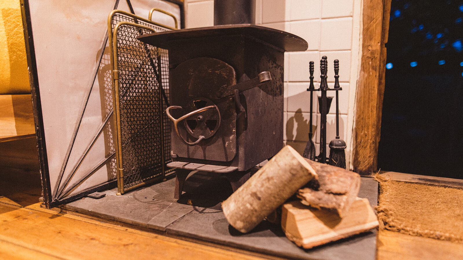 Wood-burning stove in a charming Woodcutters Cottage, logs and a metal screen nearby, set on a tiled hearth with fireplace tools in the background.