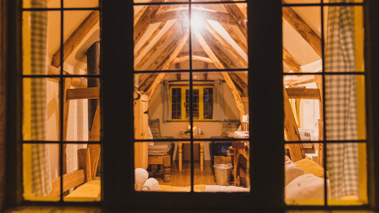 Peering through the window into this snug attic room, reminiscent of a woodcutter's cottage, you find wooden beams gracefully framing a table and chairs basked in warm lighting.