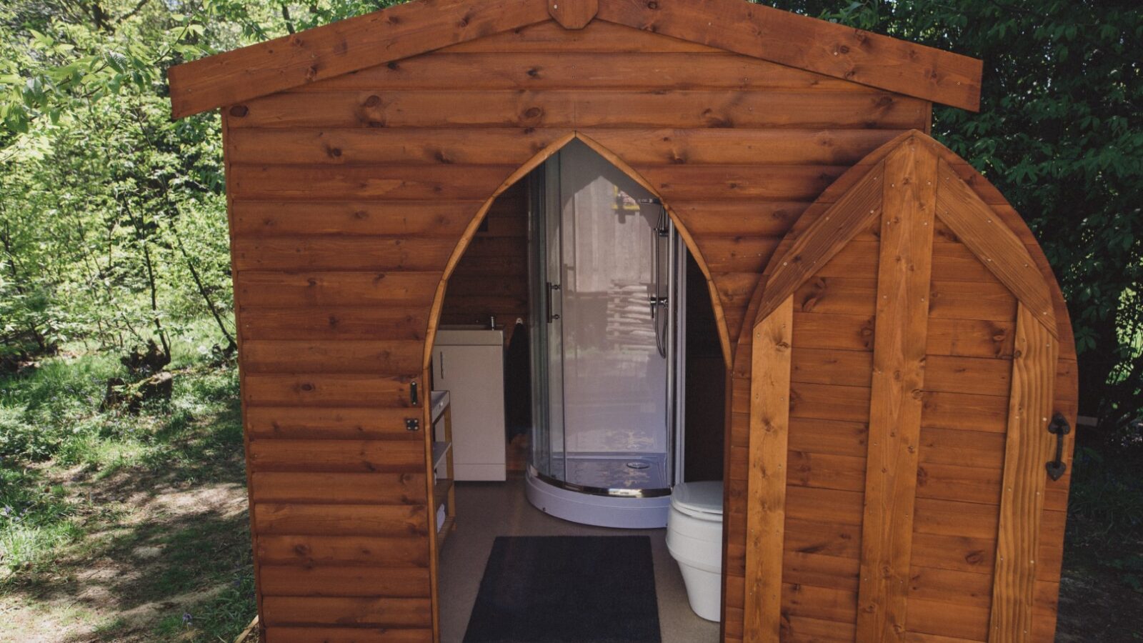 Woodcutters Cottage features a wooden cabin with a charming pointed arched door, complete with a toilet and shower inside, nestled in a serene forested area.