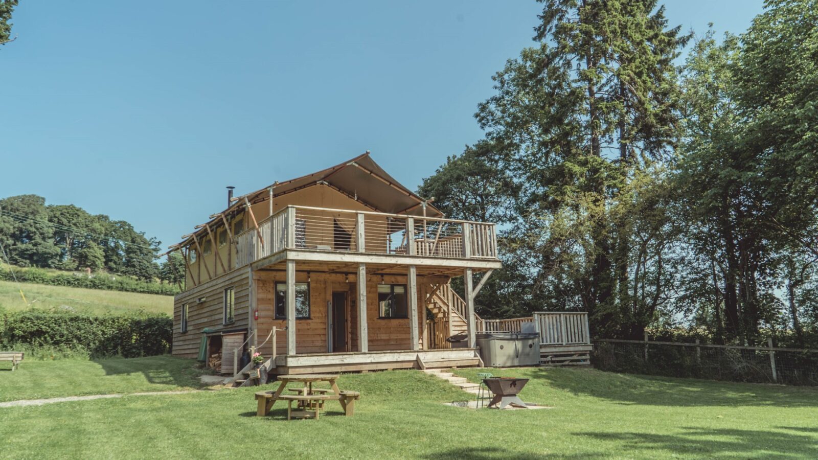 Nestled on Sweeney Farm, this two-story wooden cabin offers a touch of glamping with its large deck and stairs. Surrounded by tall trees under a clear blue sky, it features a picnic table and fire pit, making it perfect for enjoying the serene outdoors.