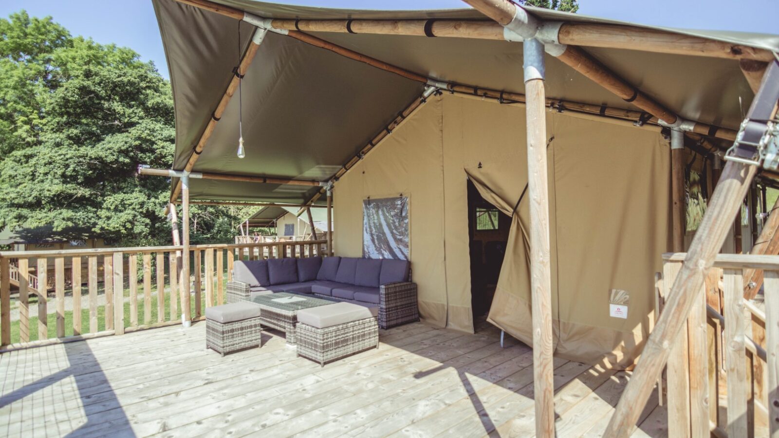 At Sweeney Farm, the glamping tent with a wooden deck is furnished with a comfortable outdoor sofa and wicker ottoman. The canvas exterior of the tent sits amid lush greenery under a clear blue sky, offering a perfect getaway.