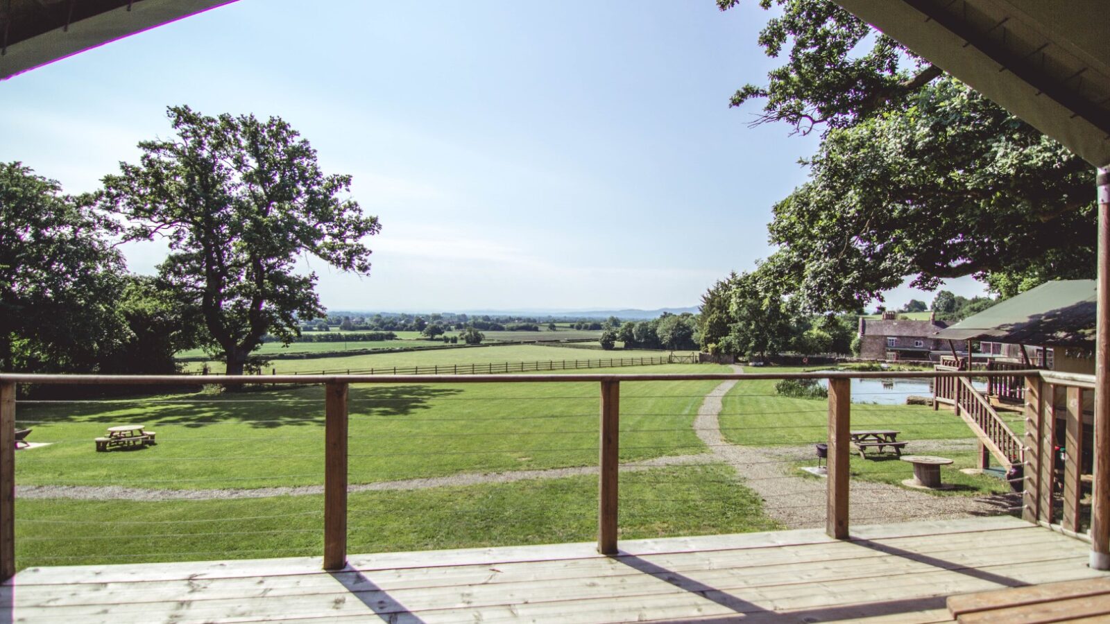 From the wooden deck of Sweeney Farm, a scenic view unfolds: a spacious, grassy field dotted with picnic benches. Large trees frame the landscape under a clear blue sky, and a path winds through the scene towards a charming glamping site in the distance.