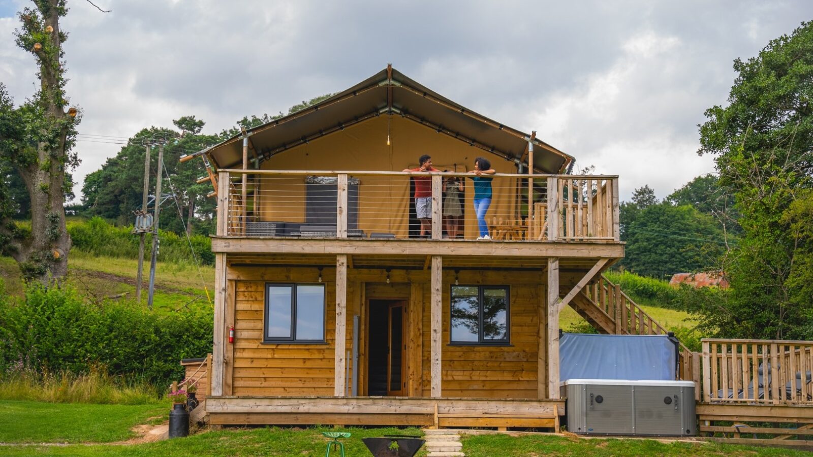 A two-story wooden cabin at Sweeney Farm boasts an upper balcony, where two people enjoy a tent-like canopy. This glamping retreat overlooks a lush grassy field with trees beneath a cloudy sky. A small, round fire pit completes the serene scene in front of the cabin.