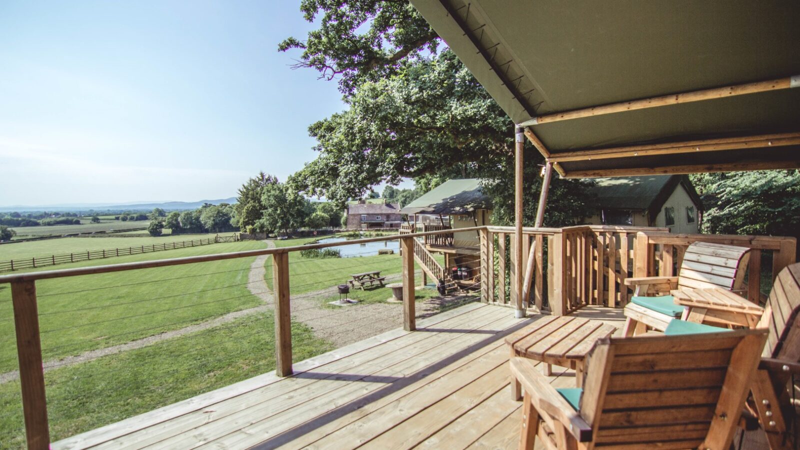 A wooden deck with chairs overlooks a grassy field and pond at Sweeney Farm, surrounded by trees. Tents hint at glamping adventures beyond a wooden fence, all under a clear blue sky.