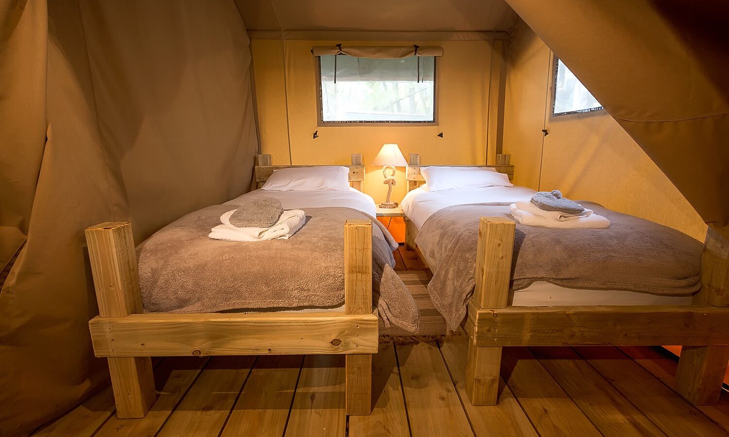 The cozy glamping tent at Sweeney Farm features two single beds with wooden frames. Each bed has neatly folded towels and a beige blanket. A small nightstand with a lamp sits between them, while soft light and tent fabric walls create a warm ambiance.