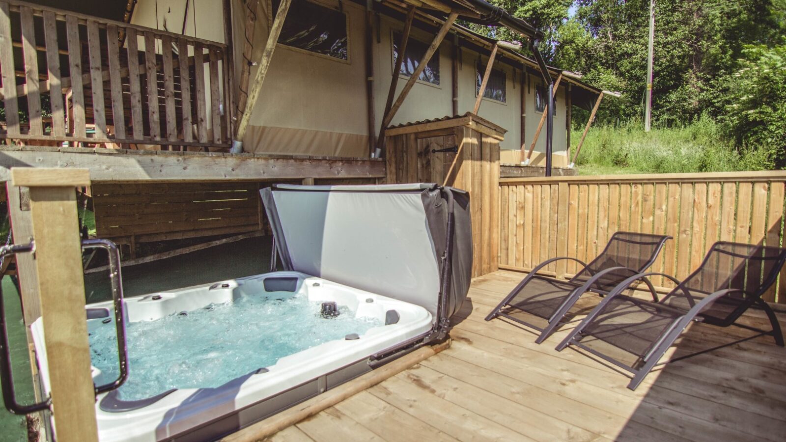 Sweeney Farm offers a serene glamping experience with an outdoor wooden deck featuring a bubbling hot tub and two black reclining chairs. Nearby, a safari-style tent cabin is nestled among lush greenery, creating the perfect tranquil retreat.