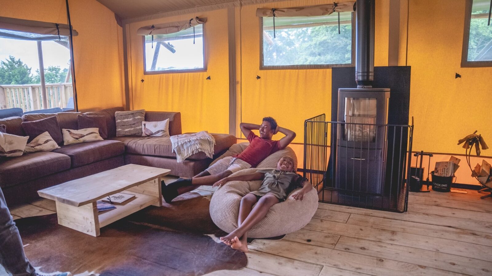 Two people are relaxing indoors on cozy seating, surrounded by rustic cabin decor at Sweeney Farm. One sits on a couch and another on a bean bag near the fireplace. Large windows provide natural light, highlighting the warm interior tones, perfect for a glamping getaway.