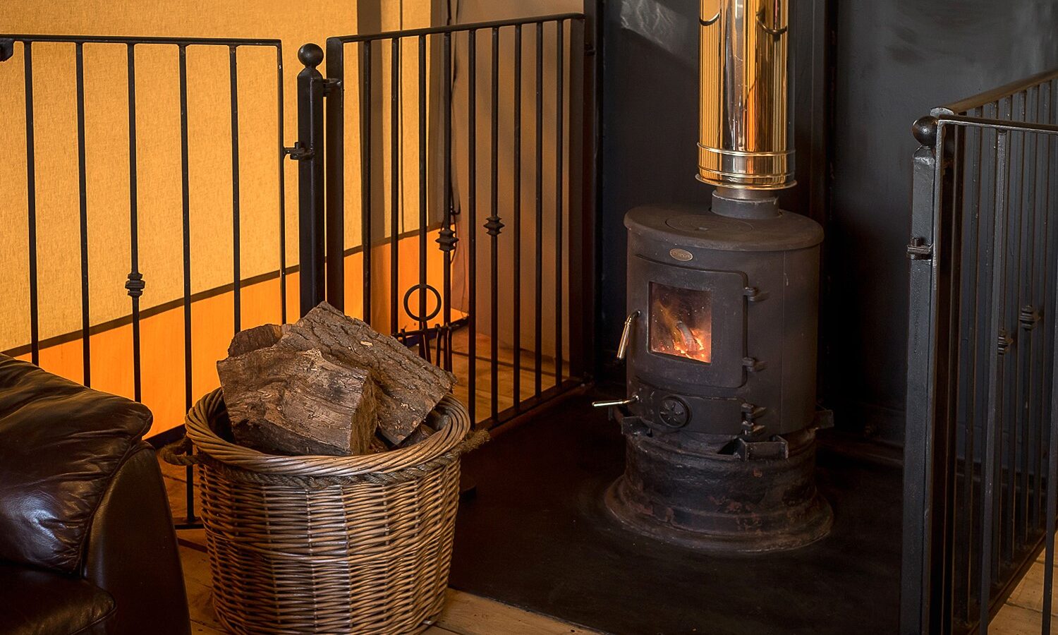 In a cozy indoor setting reminiscent of glamping at Sweeney Farm, a small wood-burning stove crackles warmly. A wicker basket brimming with firewood rests nearby, protected by a black safety guard. The inviting glow casts soft shadows on a partially visible leather sofa.