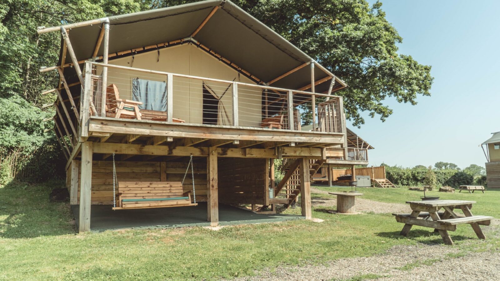 Nestled in the trees, this Sweeney Farm glamping cabin boasts a canvas roof and raised wooden structure. Enjoy the porch with its chairs and railing, or relax on the swing below. Nearby, a picnic table invites you to dine under a clear, blue sky.