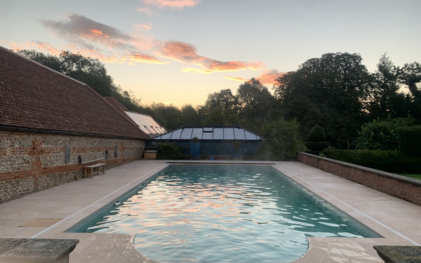 A serene outdoor swimming pool surrounded by rustic brick walls and lush greenery at sunset, located at West Lexham Treehouses. A small building peeks out under the colorful sky, adding to the tranquil charm of the West Lexham resort experience.