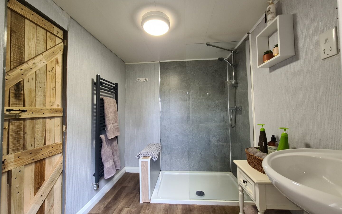 This charming bathroom, nestled within a cozy treehouse, features a shower, wooden barn door, towel rack, and a white sink with toiletries on a small table.