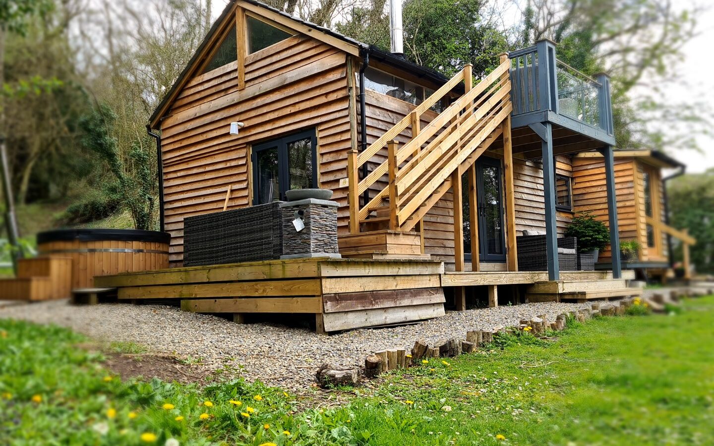 A small wooden cabin with a balcony and outdoor seating in a grassy area surrounded by trees, perfect for star gazing. Relax in the hot tub while enjoying the serene view, creating an enchanting treehouse-like escape under the stars.