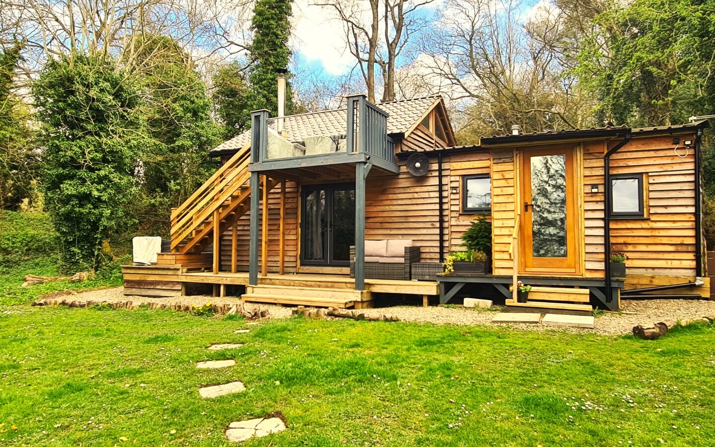 A rustic wooden cabin with a porch and loft, nestled like a star gazing treehouse amidst a green lawn and surrounded by tall trees on a clear day.