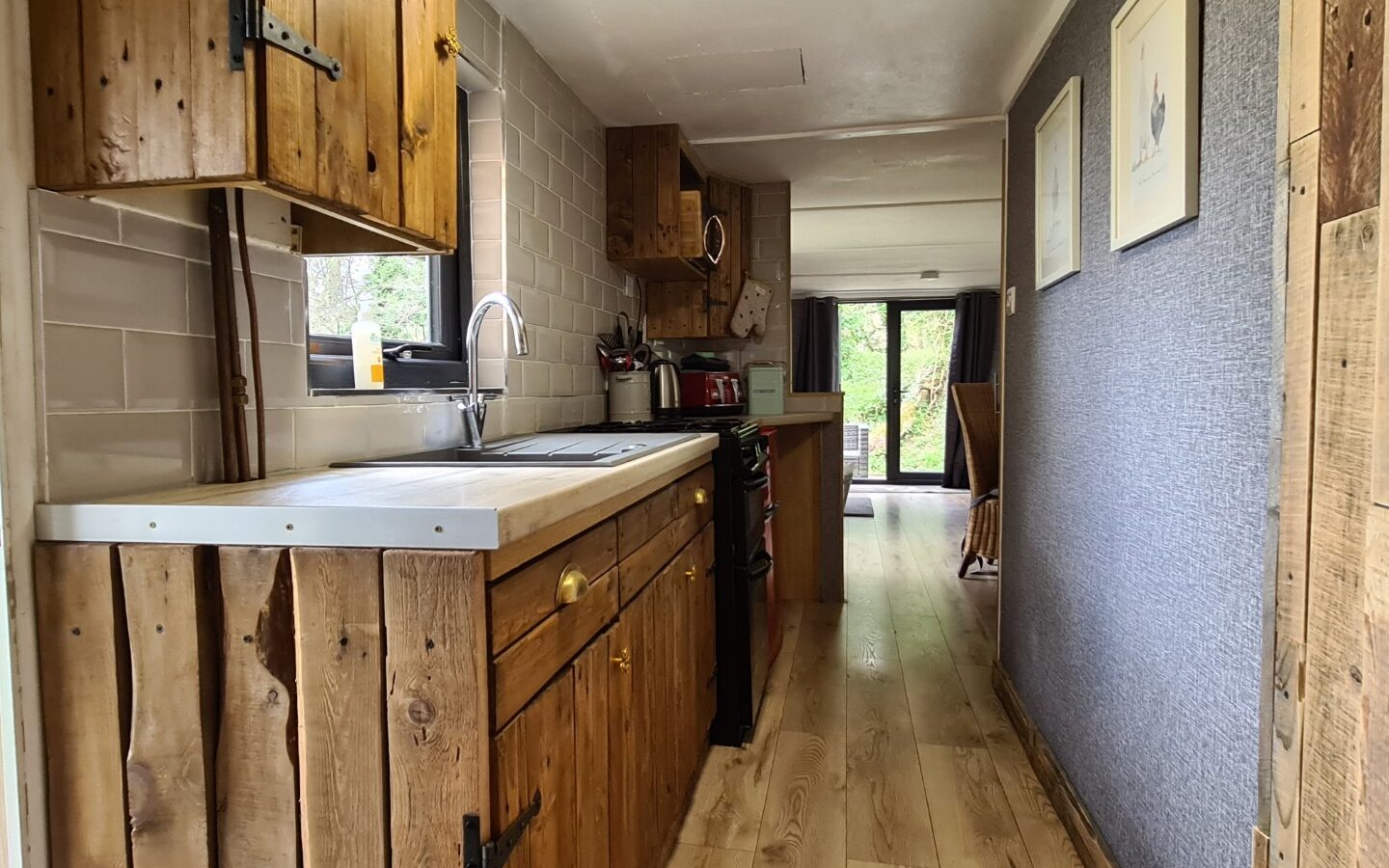 A narrow kitchen with wooden cabinets, a sink, stove, and tiled backsplash feels like the cozy heart of a treehouse. A hallway leads to a room filled with natural light.