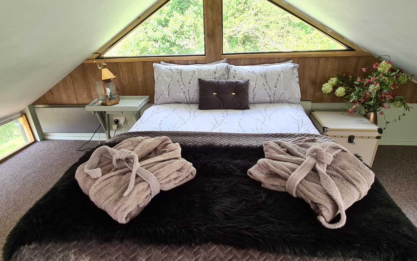 Cozy attic bedroom reminiscent of a treehouse, featuring a double bed, two folded bathrobes, triangular windows perfect for stargazing, bedside tables, and decorative plants.