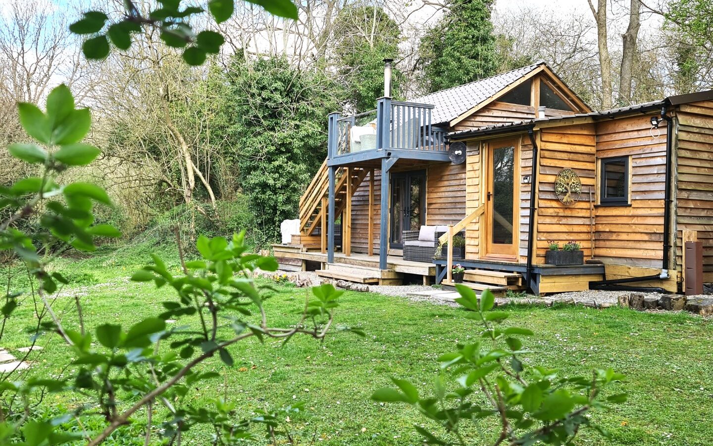 A wooden cabin reminiscent of a treehouse features a small deck ideal for star gazing, nestled amidst lush greenery and trees on a grassy plot of land.